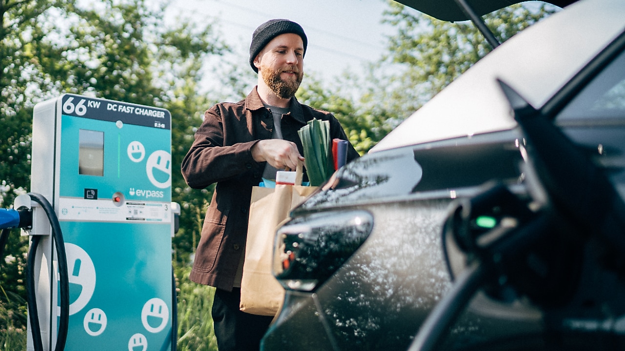 Un homme range des sachets de courses dans le coffre de sa voiture électrique. La voiture est raccordée et en charge, la borne de recharge est visible en arrière-plan.